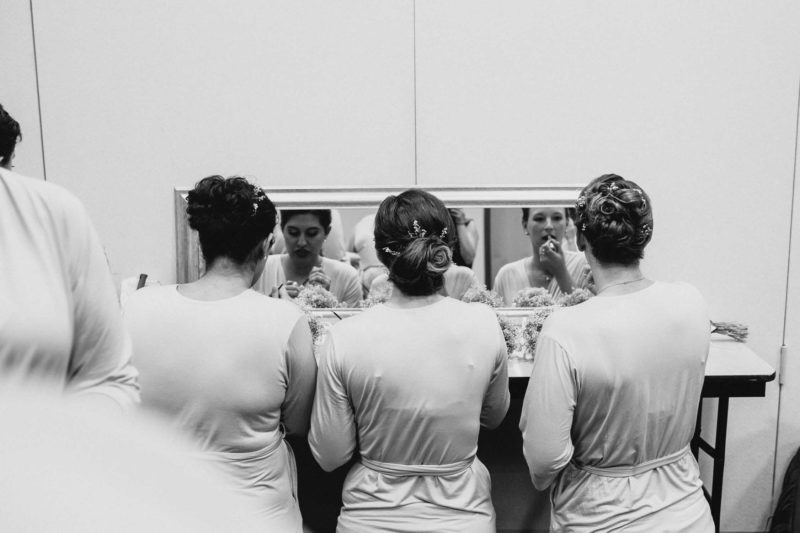 Black and white photo or three women sitting down facing a mirror. We see their backs and the reflection of them putting on makeup.