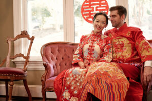 A couple dressed in traditional Chinese wedding attire sit on a pink sofa. The woman is looking at the camera, the man is looking at his wife.