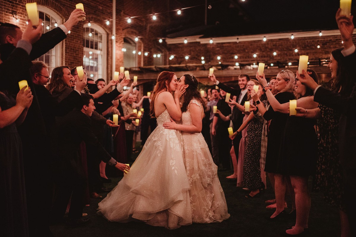 Two brides kiss while surrounded by guests holding LED candles. There are market lights above them, and it is nighttime.