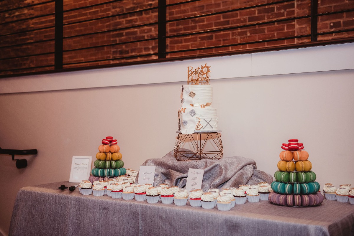 A dessert table featuring a two tier wedding cake, cupcakes, and two rainbow macaron towers
