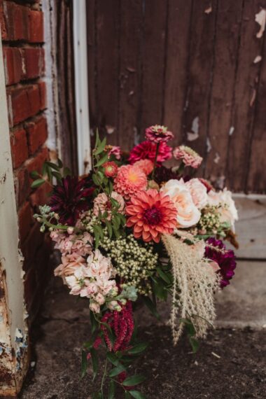 A colorful bouquet with oranges, reds, corals, and purples sits on the ground next to a brick wall.