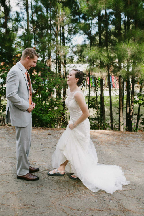 A white groom wearing a light grey suit and burnt orange vest smiles as he watched his bride reveal that she is wearing chacos under her wedding gown.
