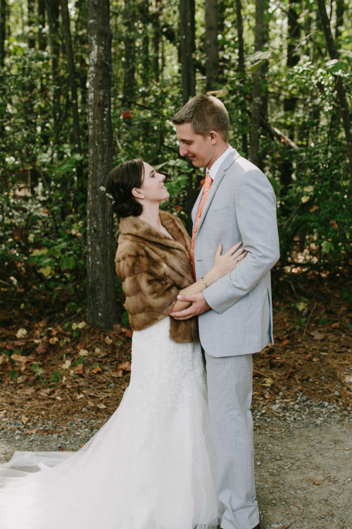 A white bride in a white gown and brown fur coat holds her groom, who is wearing a light grey suit with a burnt orange vest. They are looking at each other.