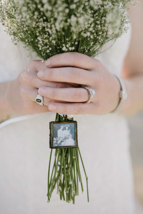 Close up on white hands holding a bouquet of babies breath. Attached to the bouquet is a miniature picture frame showing a black and white photo of a bride and groom.