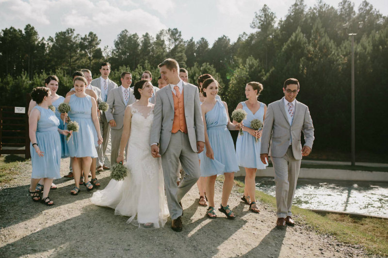 A bride and groom look at each other smiling, holding hands while walking slightly ahead of their wedding party. The party is wearing light blue and light grey.