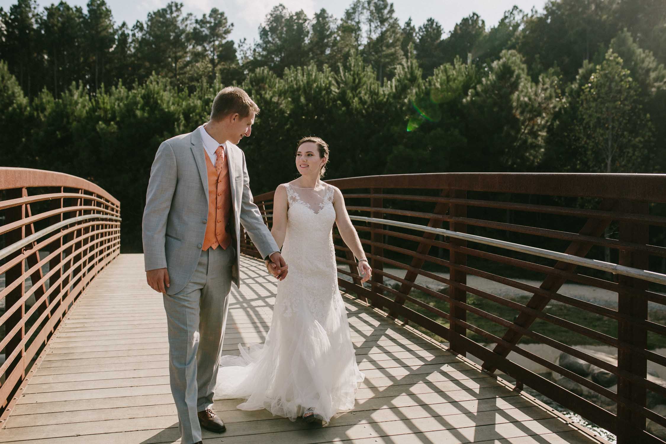 A groom in light grey and bride in white look at each other while walking toward us over a wooden bridge.