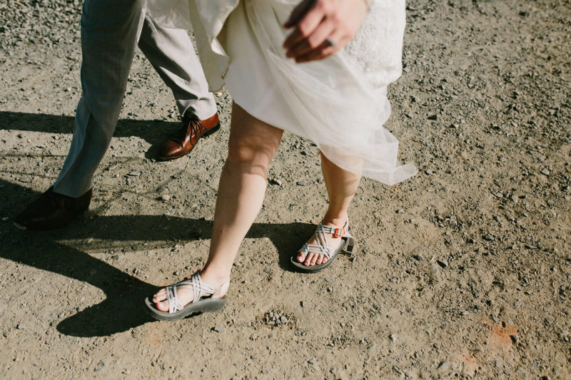 Close up on two pairs of feet walking. One person wears light grey pants and brown dress shoes, the other wears a white wedding dress and light colored Chaco sandals.