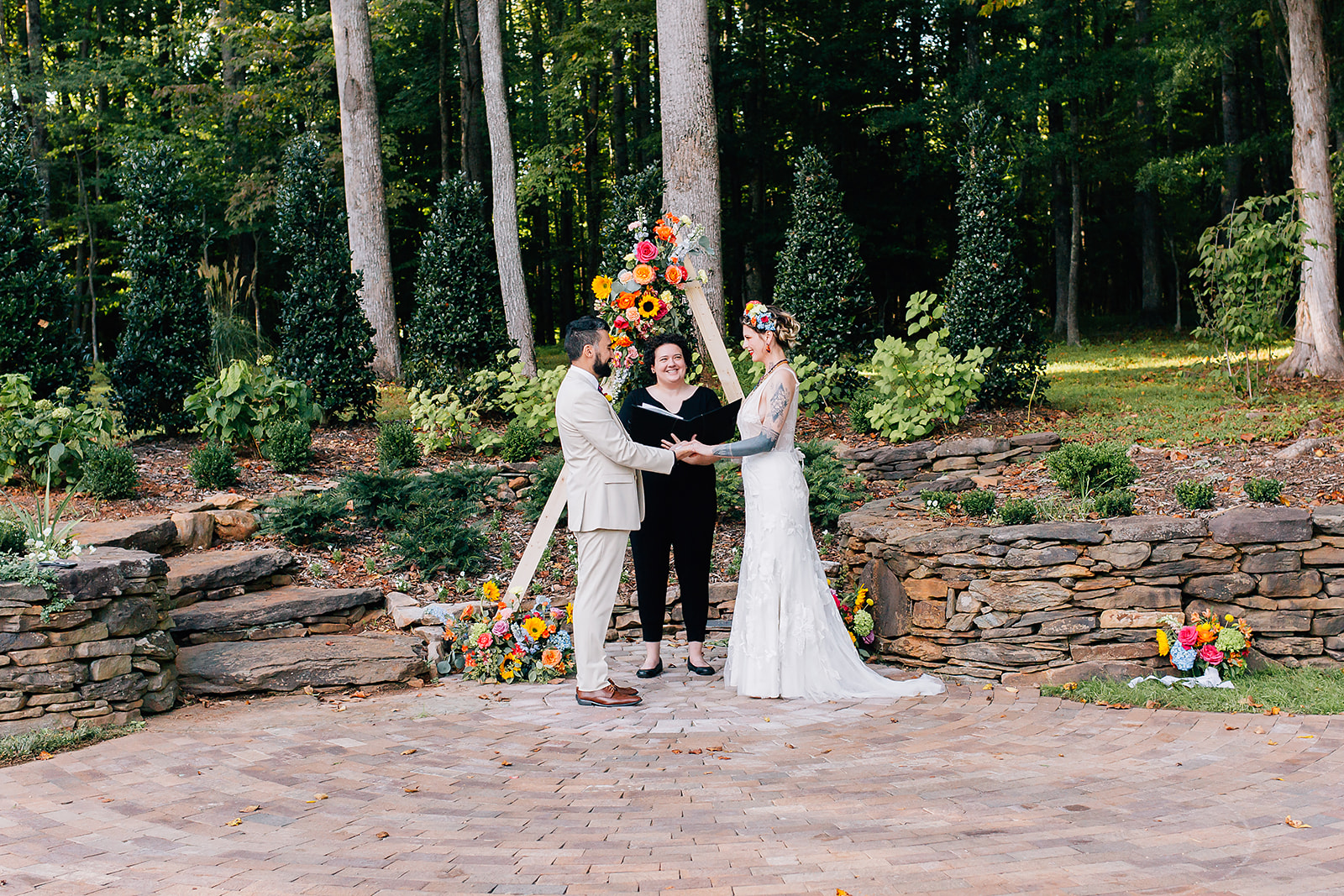 A groom and bride stand in front of an officiant, all three of them in front of a wooden triangular arbor decorated in bright flowers. 