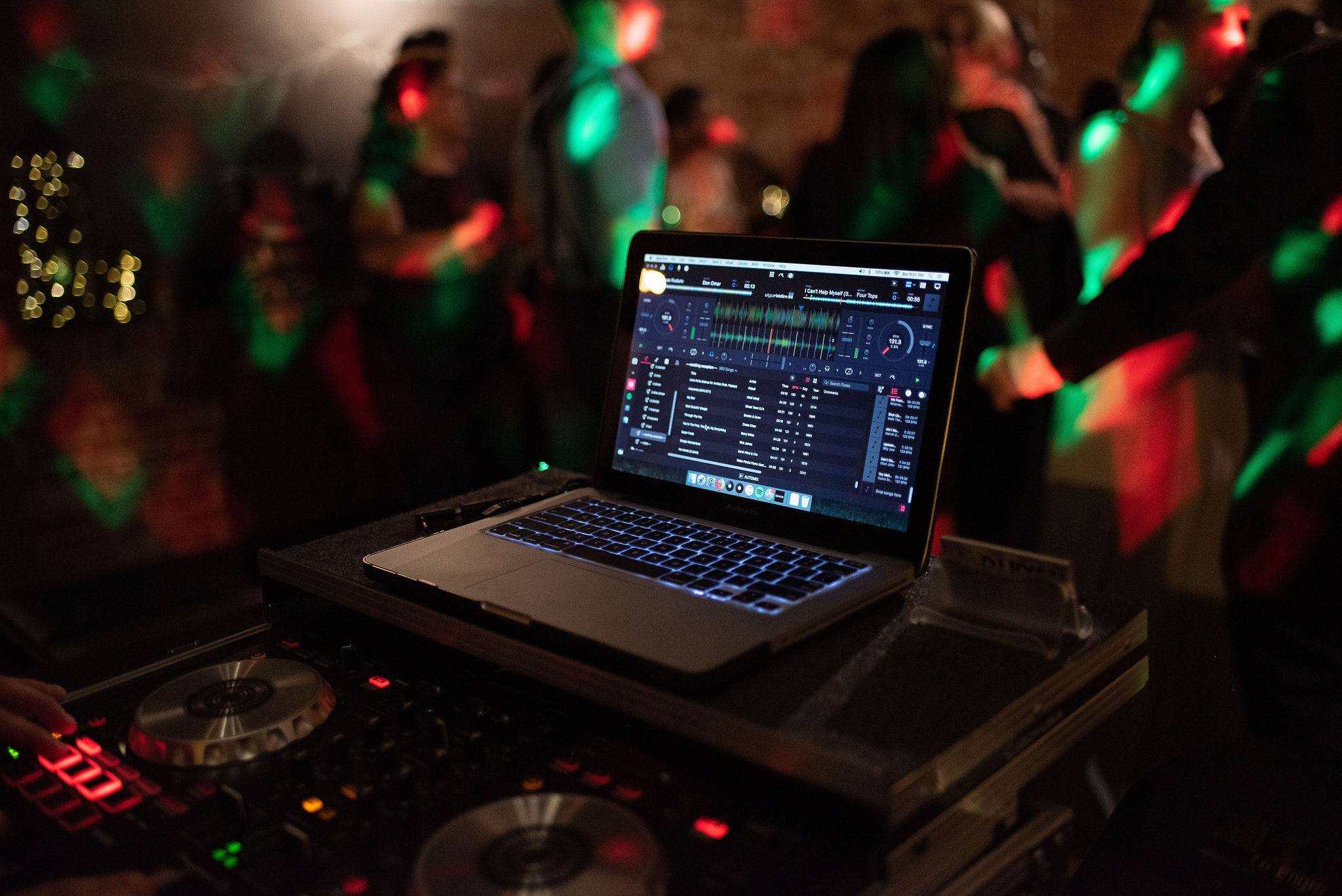 Close up of a laptop sitting on a DJ turn table. Out of focus in the background is a dance floor with colorful lighting. 