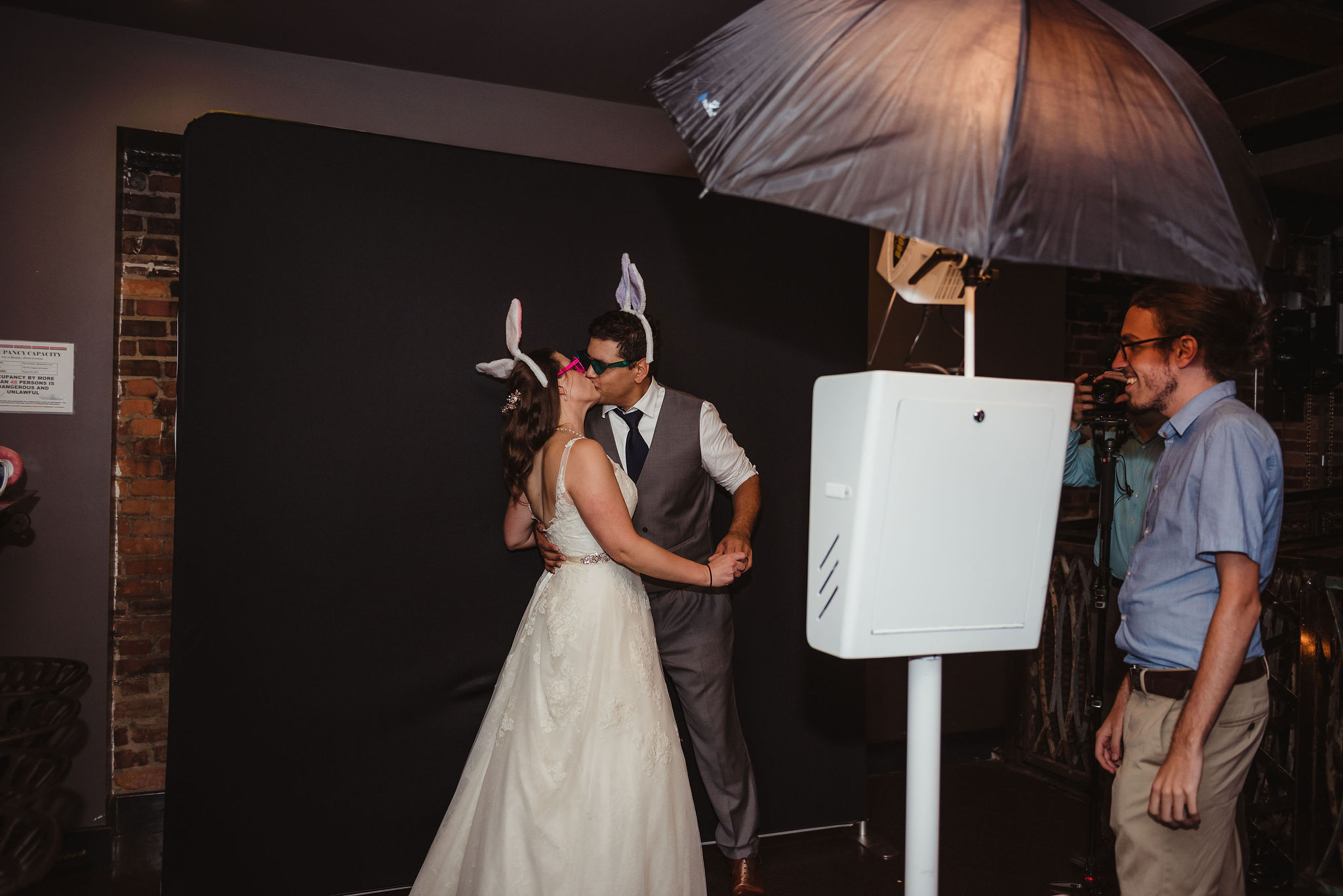 A bride and groom kiss in a photobooth. They are wearing bunny ears, and there is a black backdrop. 
