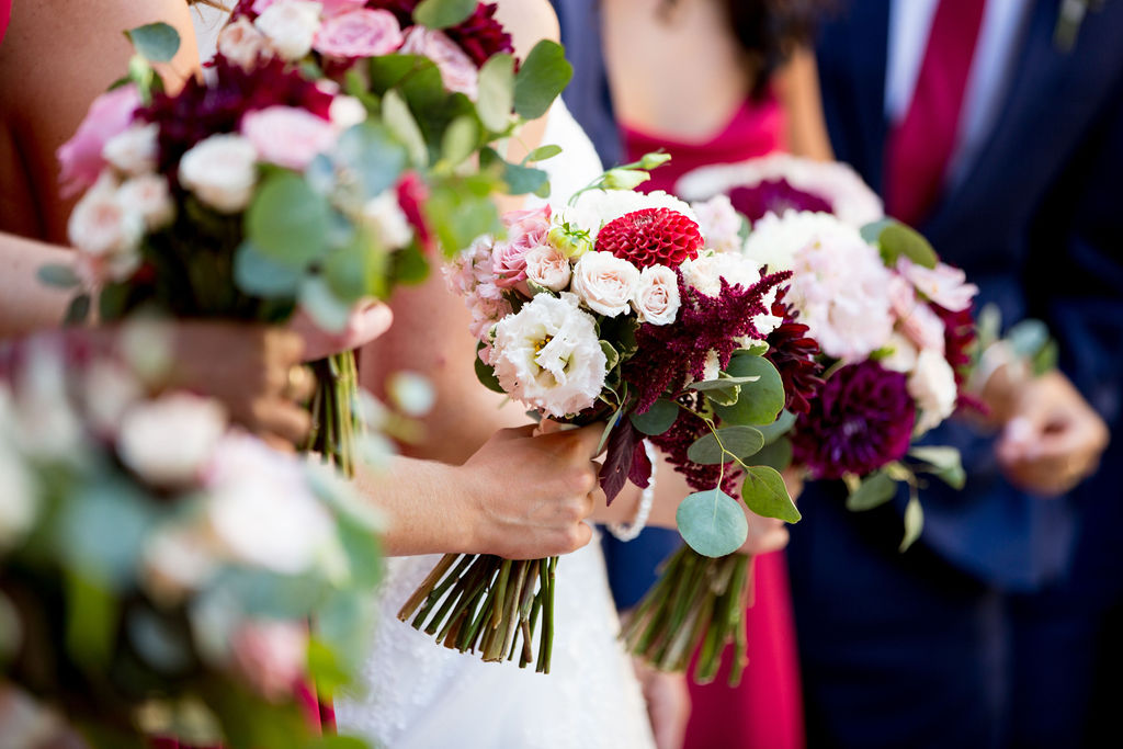 close up on a group of hands holding bouquets. The flowers in the bouquets are white, burgundy, and blush.