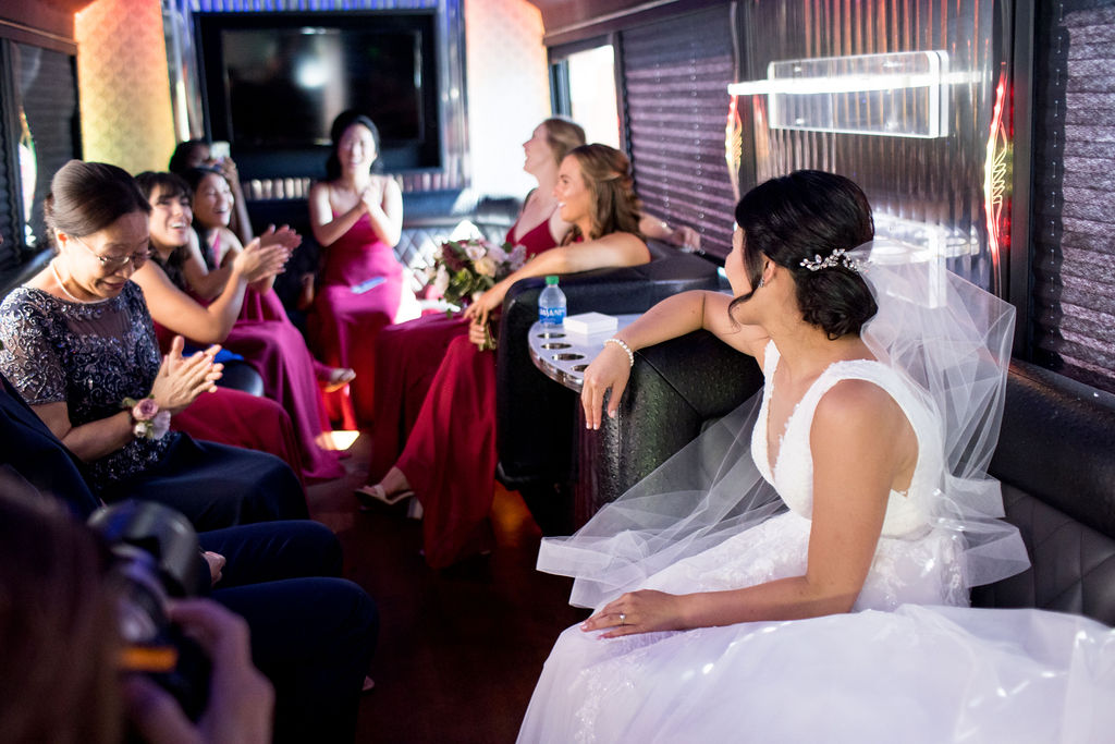 A bride sits on a party bus facing her wedding party and mom. 