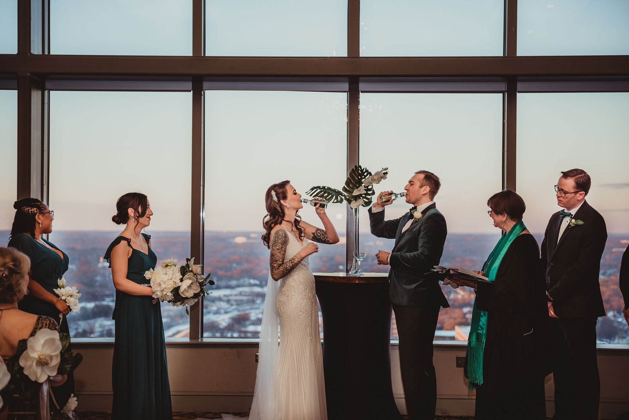 A bride and groom drink together while their wedding party watches.