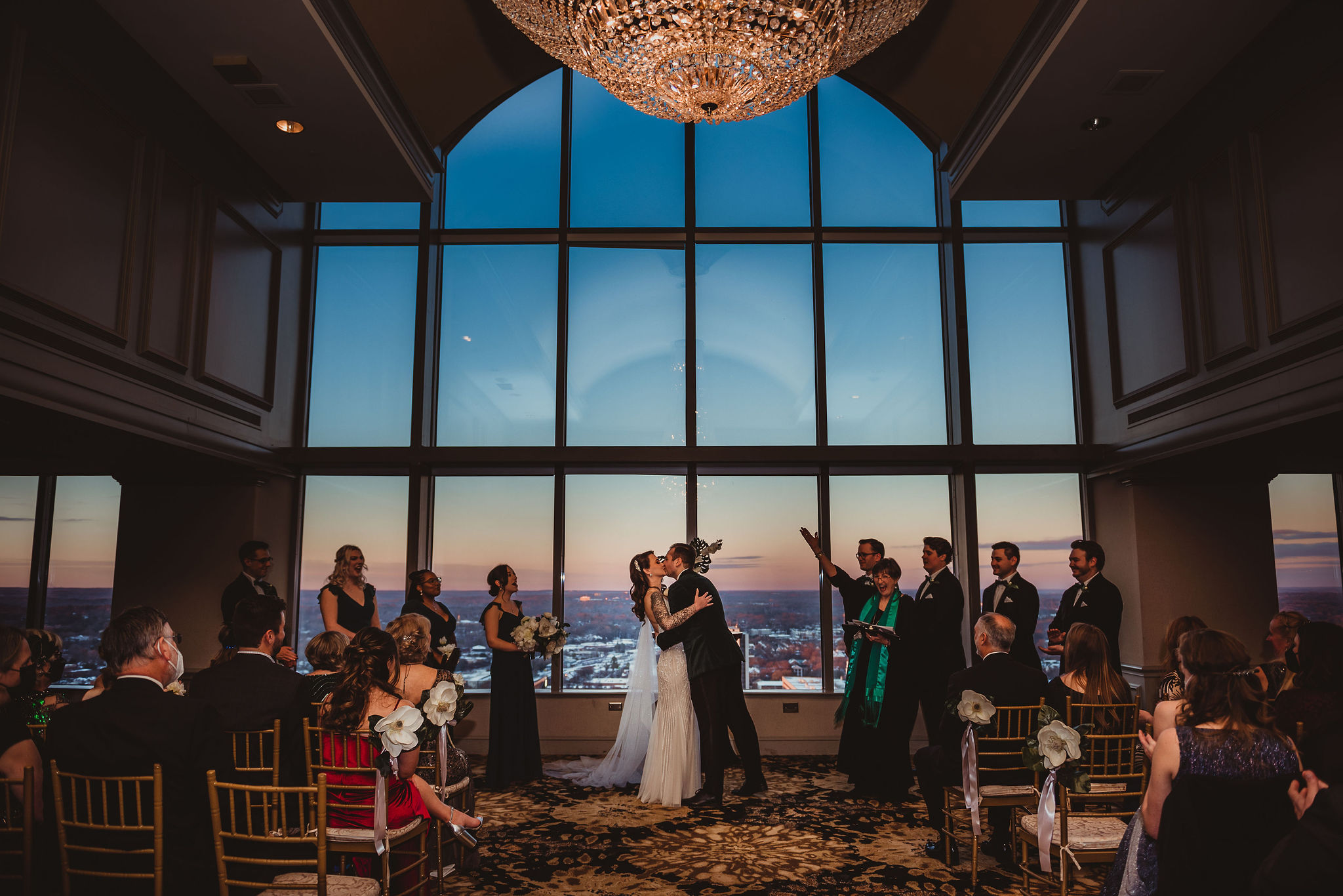 A bride and groom share their first kiss as husband and wife at the end of their ceremony, in front of large windows showing a view of a city.