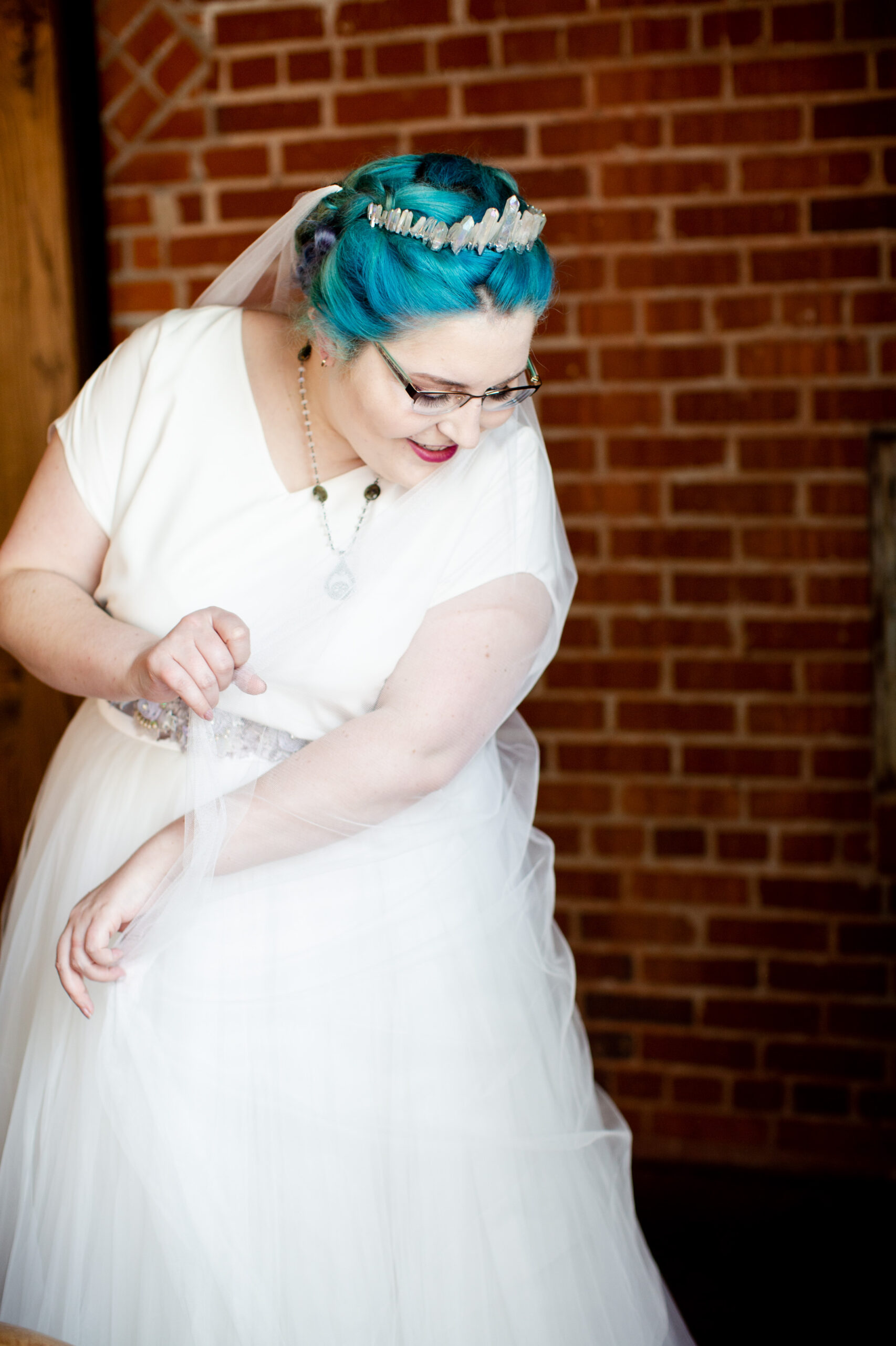 A bride with bright electric blue hair stand holding her veil over her left arm as she looking down toward the floor. 