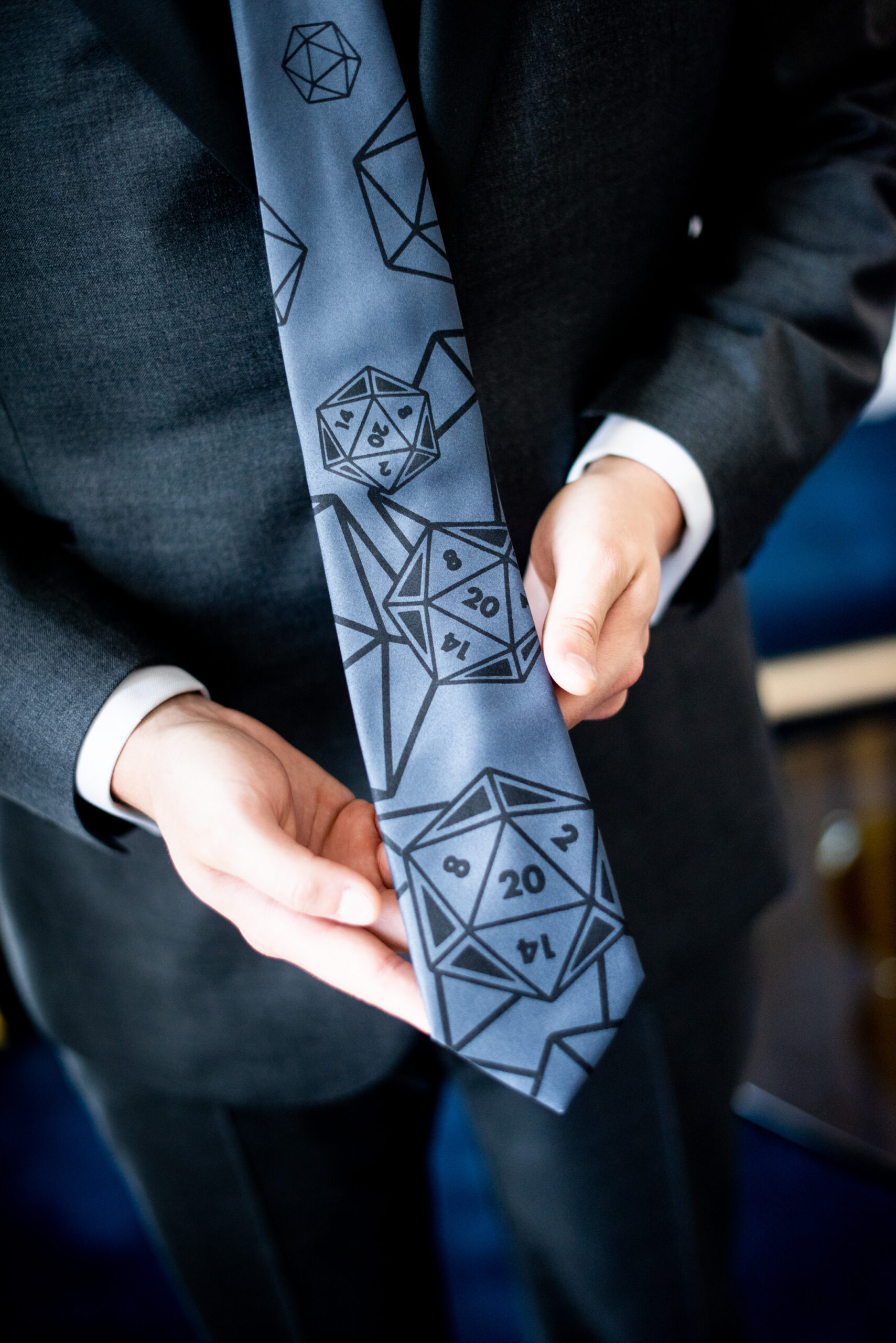 A groom in a dark grey suit holds out his blue tie for us to see. On the tie are outlines of 20-sided dice.