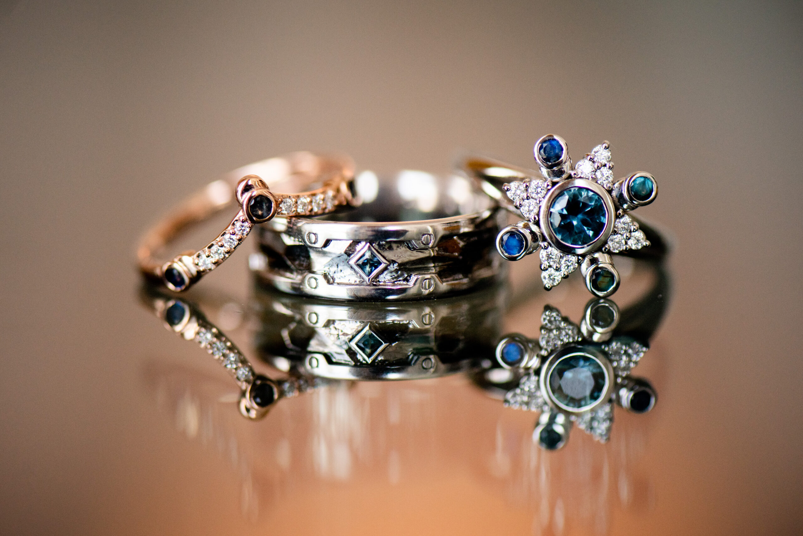 Three white gold wedding rings sit on a table. The rings are clearly custom made, with a variety of clear and blue stones and a star shape.