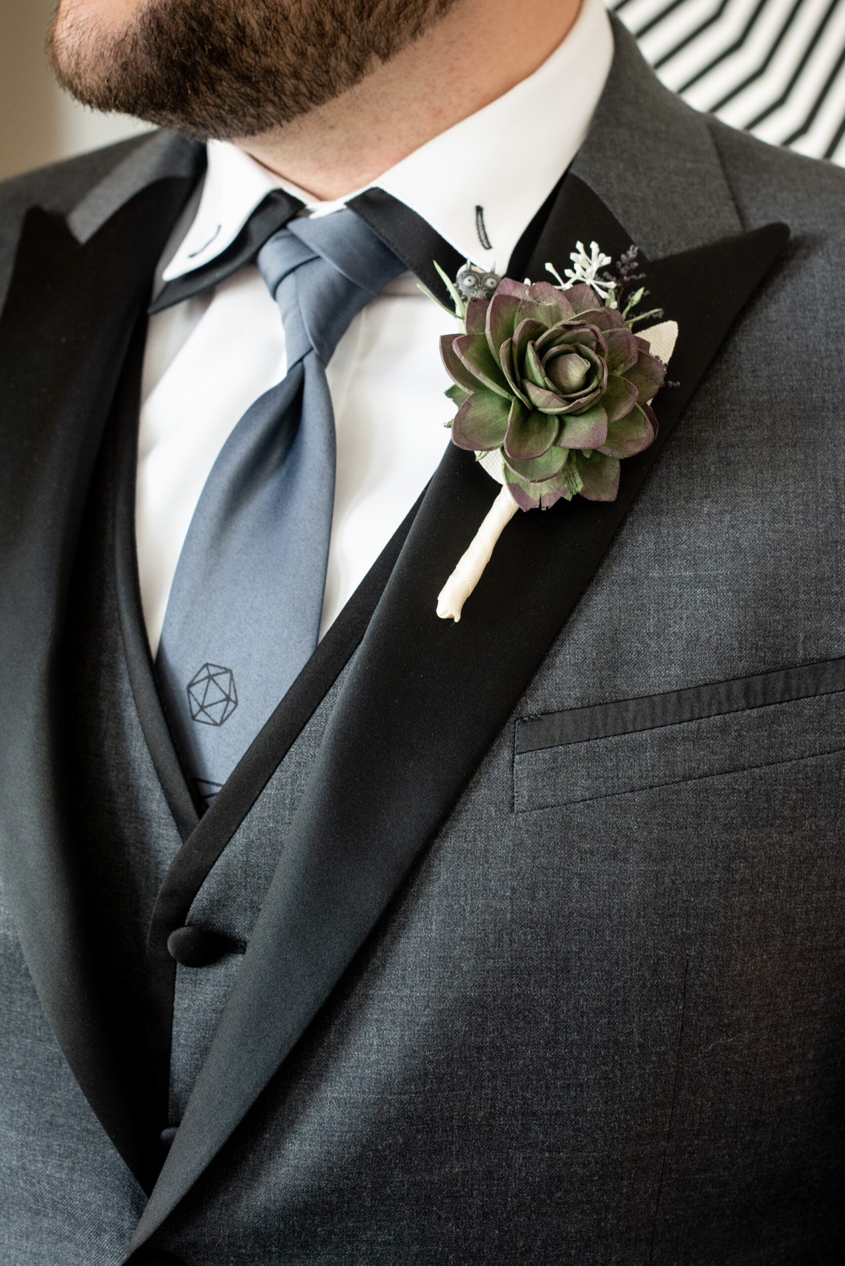 Close up on the chest of a groom dressed in a dark grey suit. The suit is buttoned, but we can see a white shirt and matching dark grey vest underneath, along with a blue tie. He also wears a succulent boutoinere.