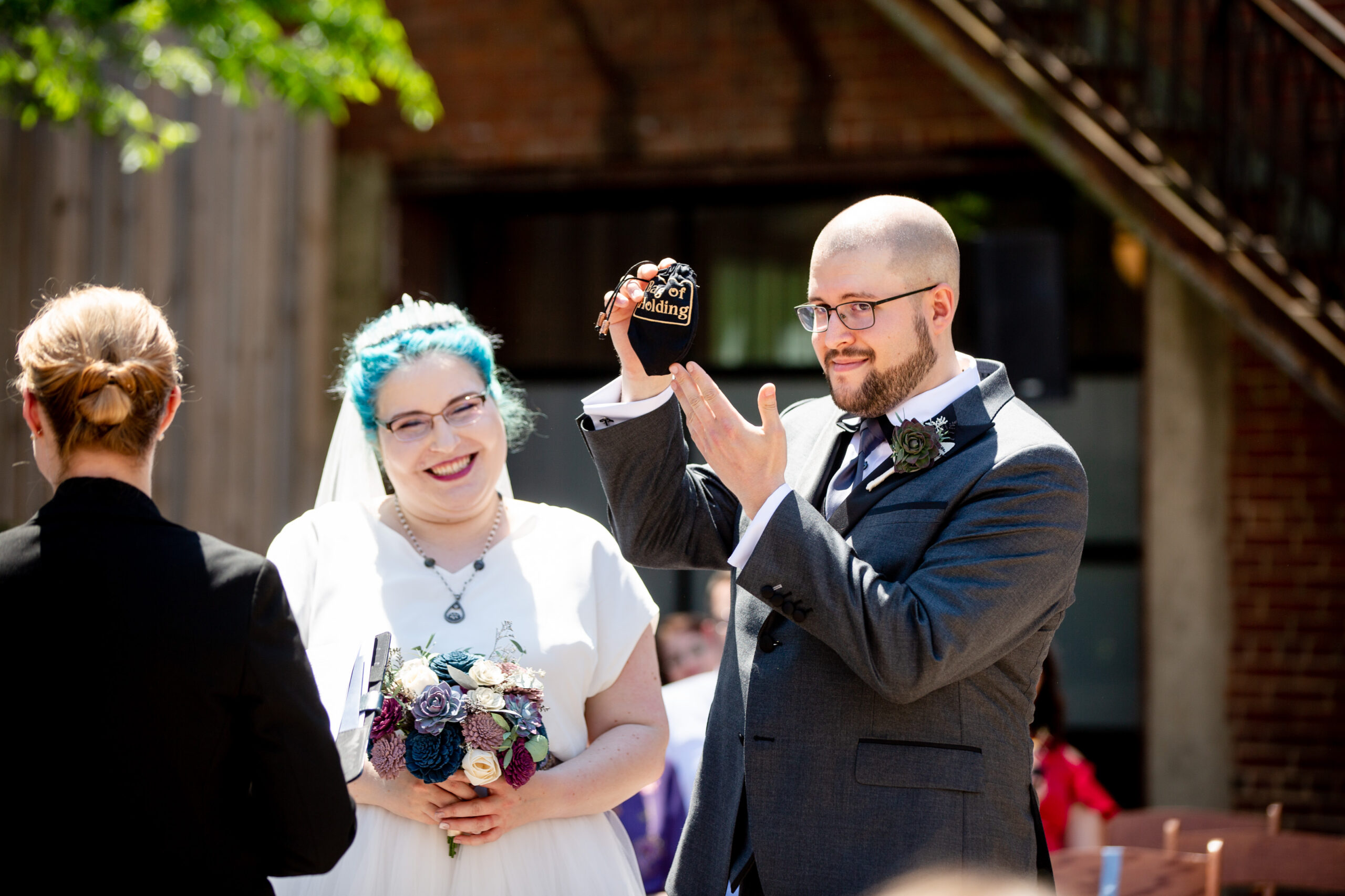 A groom holds a tiny black bag with one hand while gesturing to it with the other. His bride stands smiling nearby, holding a colorful bouquet of paper flowers. 