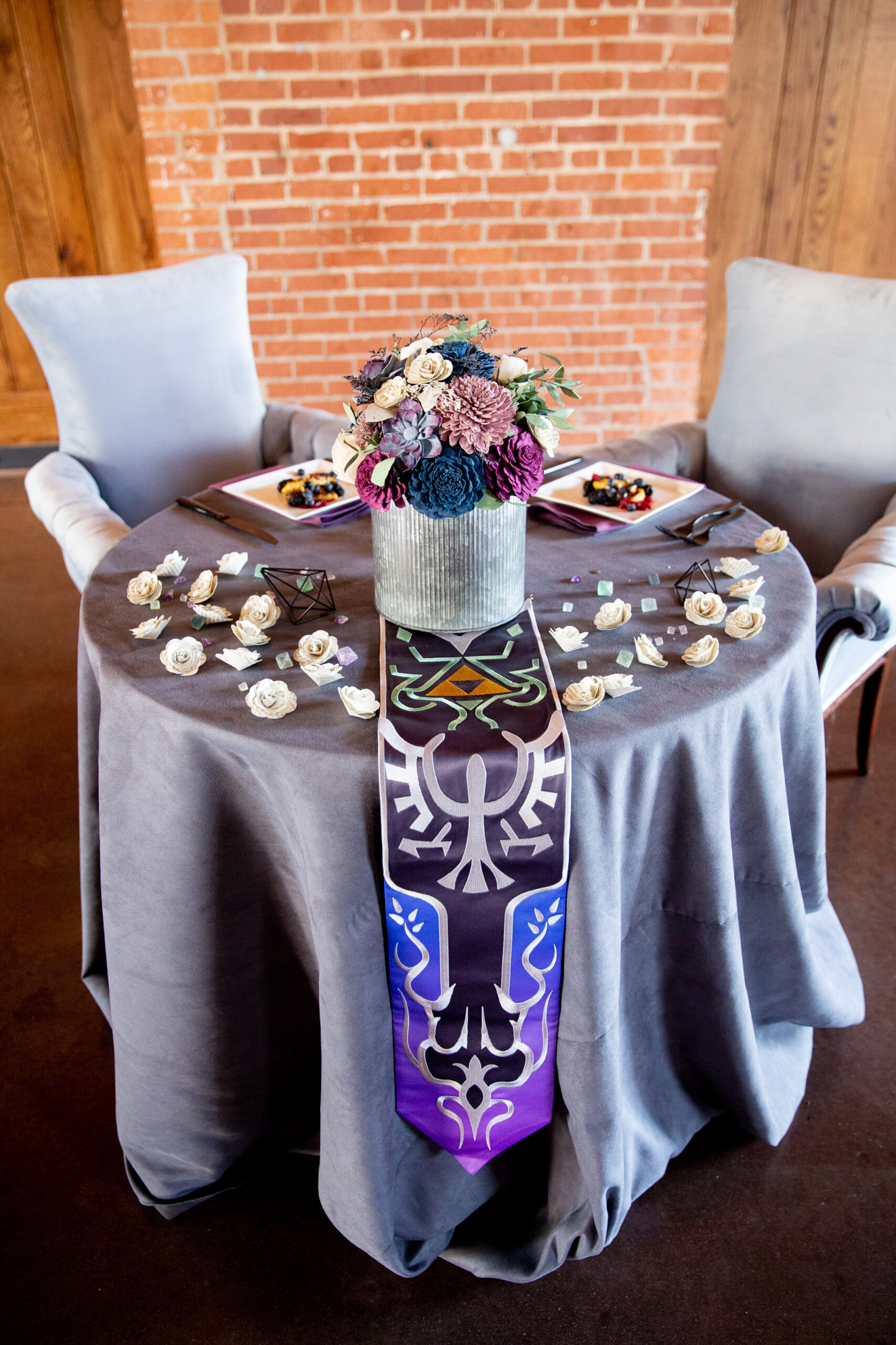 A small round sweetheart table is decorarted with a adark grey velvet cloth and topped with book page flowers. At the center is a metal vase holding a bouquet of purple and blue wood flowers. 