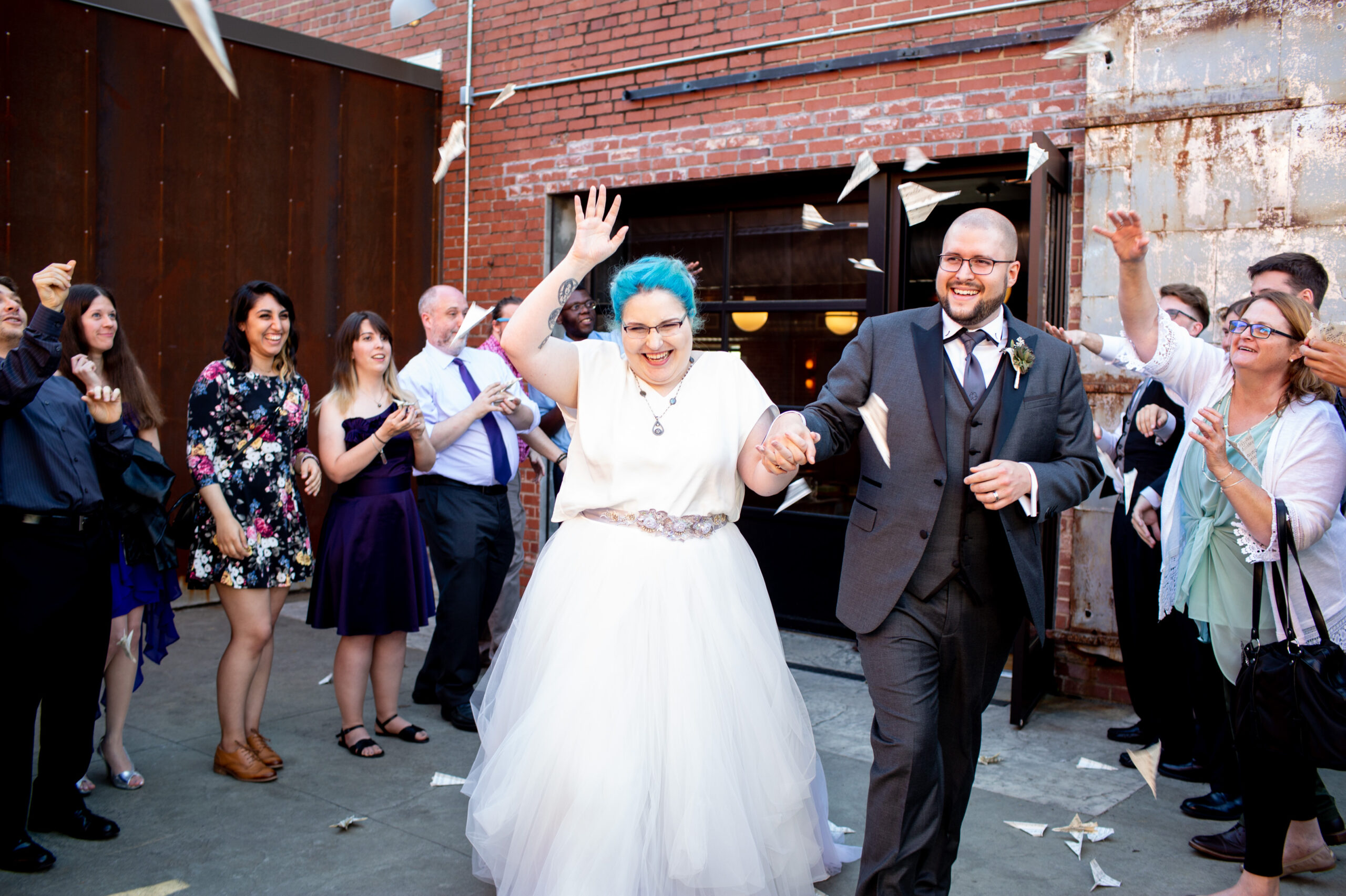 A bride with electric blue hair and a groom in a dark suit walk toward us arm in arm, as guests throw paper airplanes around them. 