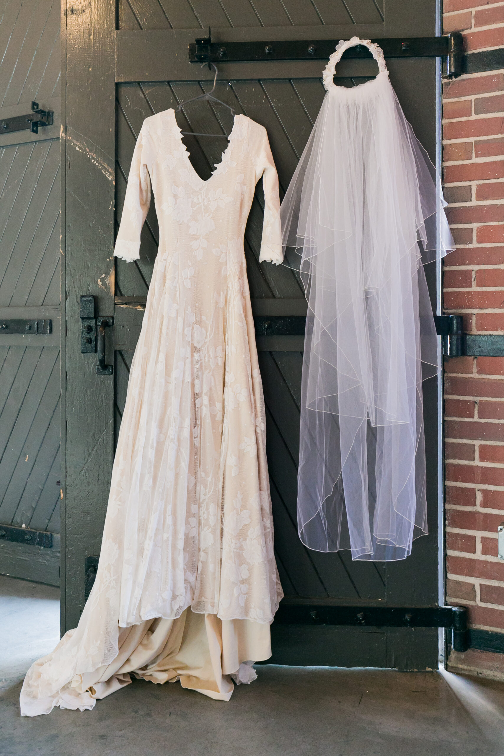 A white wedding gown with three quarter length sleeves hangs next to a white veil from a dark wooden door. 