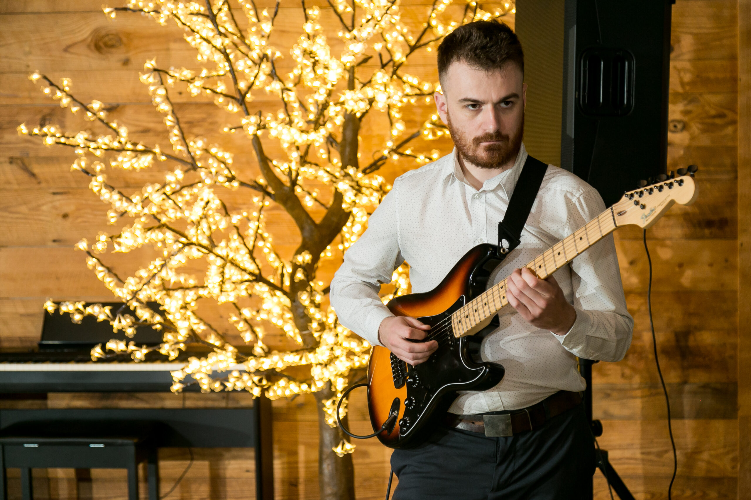A man plays an electric guitar while standing in from of a light up tree.